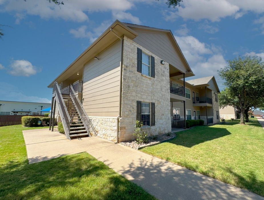 Riviera two-story beige apartment building with stone accents, exterior stairs, and a well-kept green lawn.