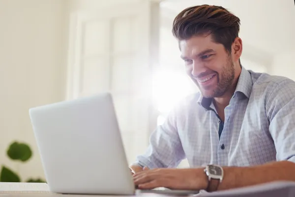 A man smiling while sitting at a table or desk looking at a laptop
