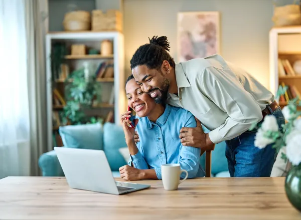 Husband hugging his wife or girlfriend while she talks on the phone smiling and looking at a laptop