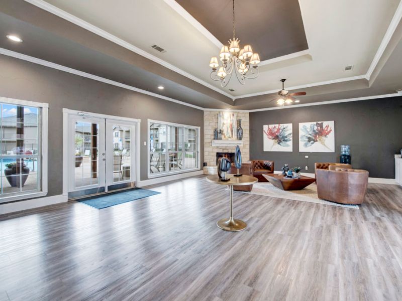 Riviera Apartments in Waco, TX community clubhouse with a cozy lounge space, fireplace, hardwood-style plank flooring, and lots of windows and natural light looking out to the pool and sun deck